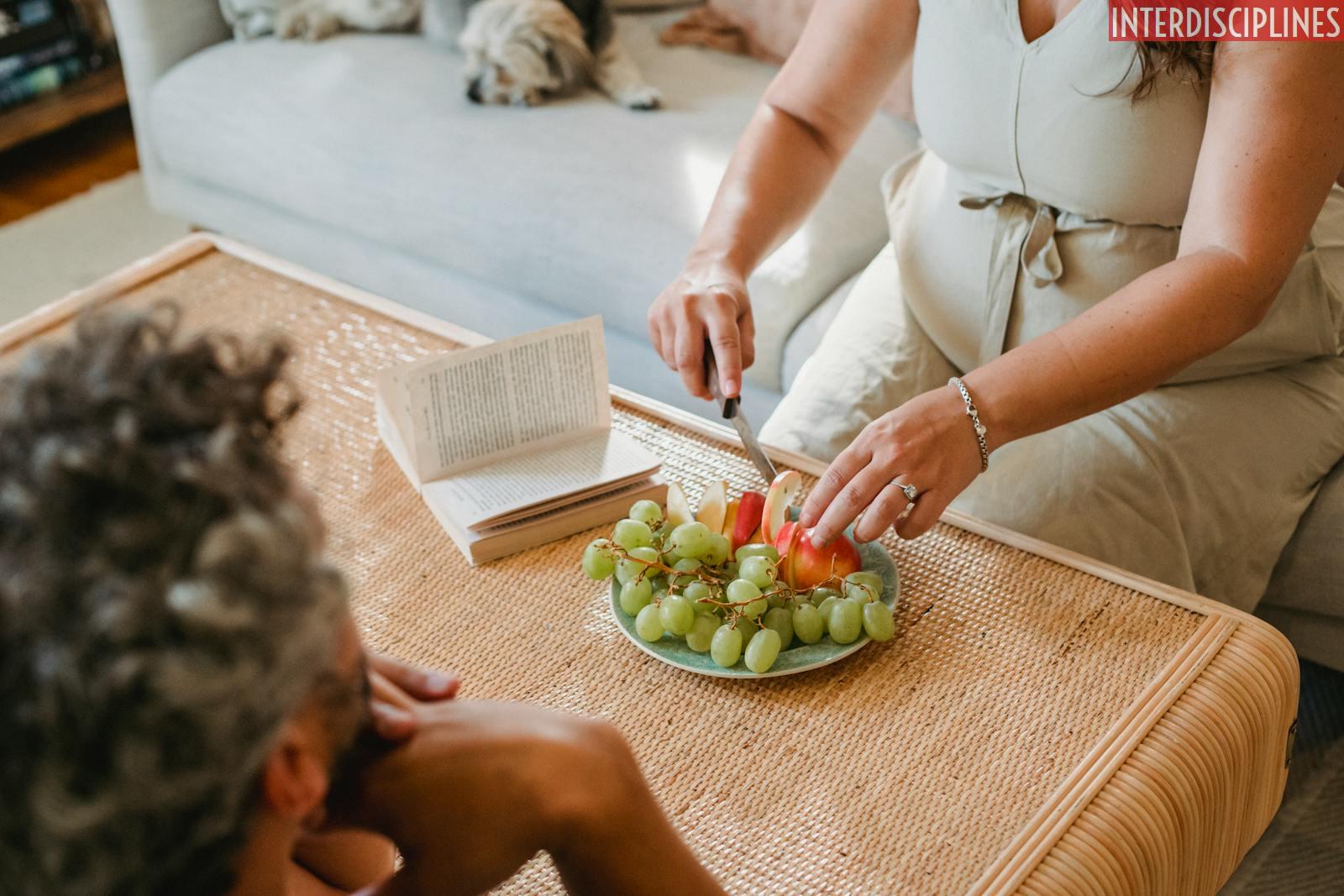 Faceless pregnant female in dress cutting apples with knife to share with husband resting on sofa